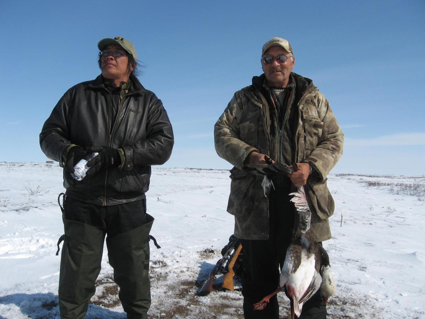 Two men stand with the duck they hunted. There is snow on the ground.