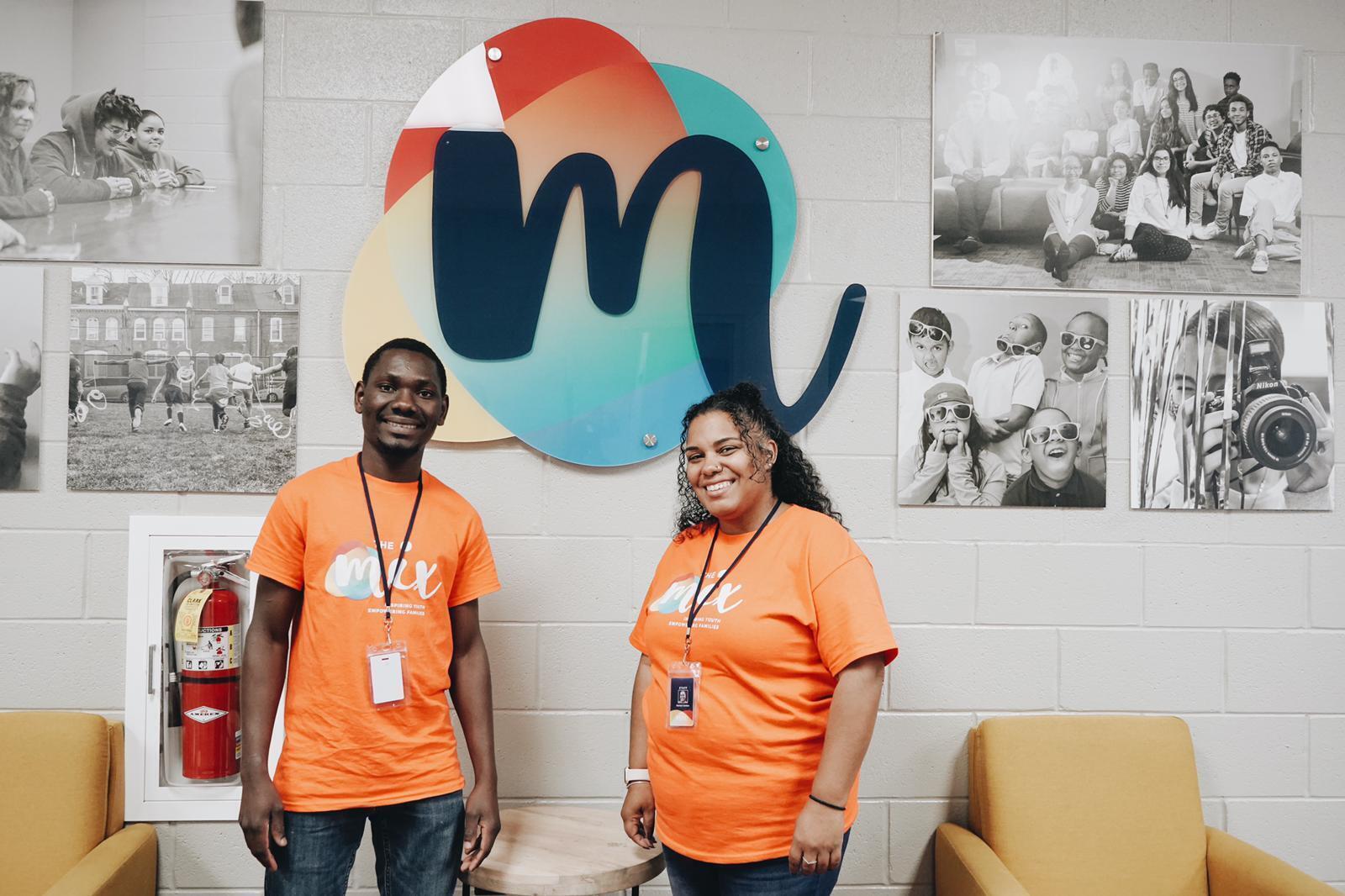 A young man and woman wearing orange t-shirts stand in front of a wall where black and white photos hang as well as a large "M" logo.
