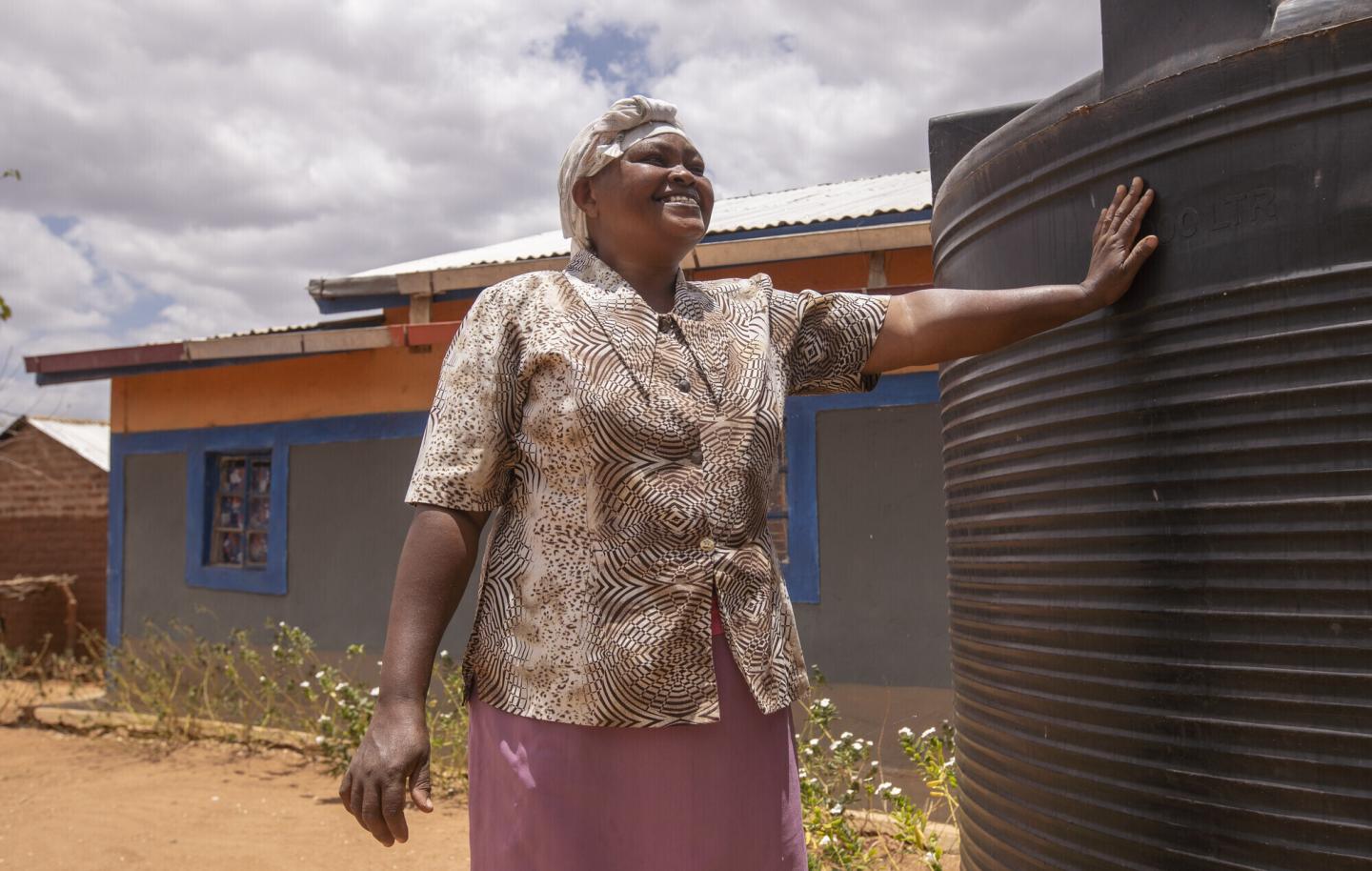 A Kenyan woman stands with her arm is outstretched, touching a rain barrel.