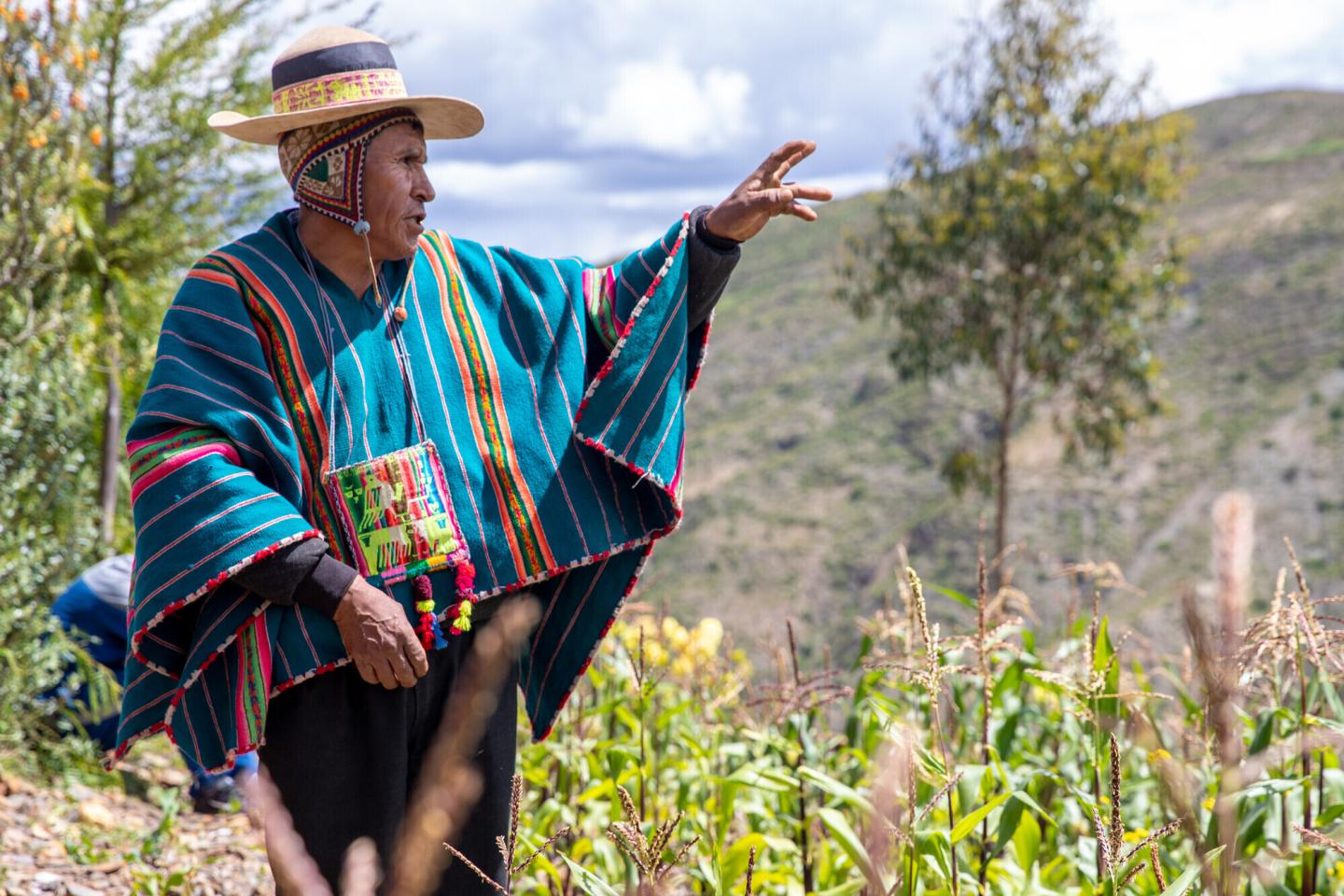 An Indigenous Bolivian man stands in a field and points to something off on the right. He is wearing a blue poncho and a hat.