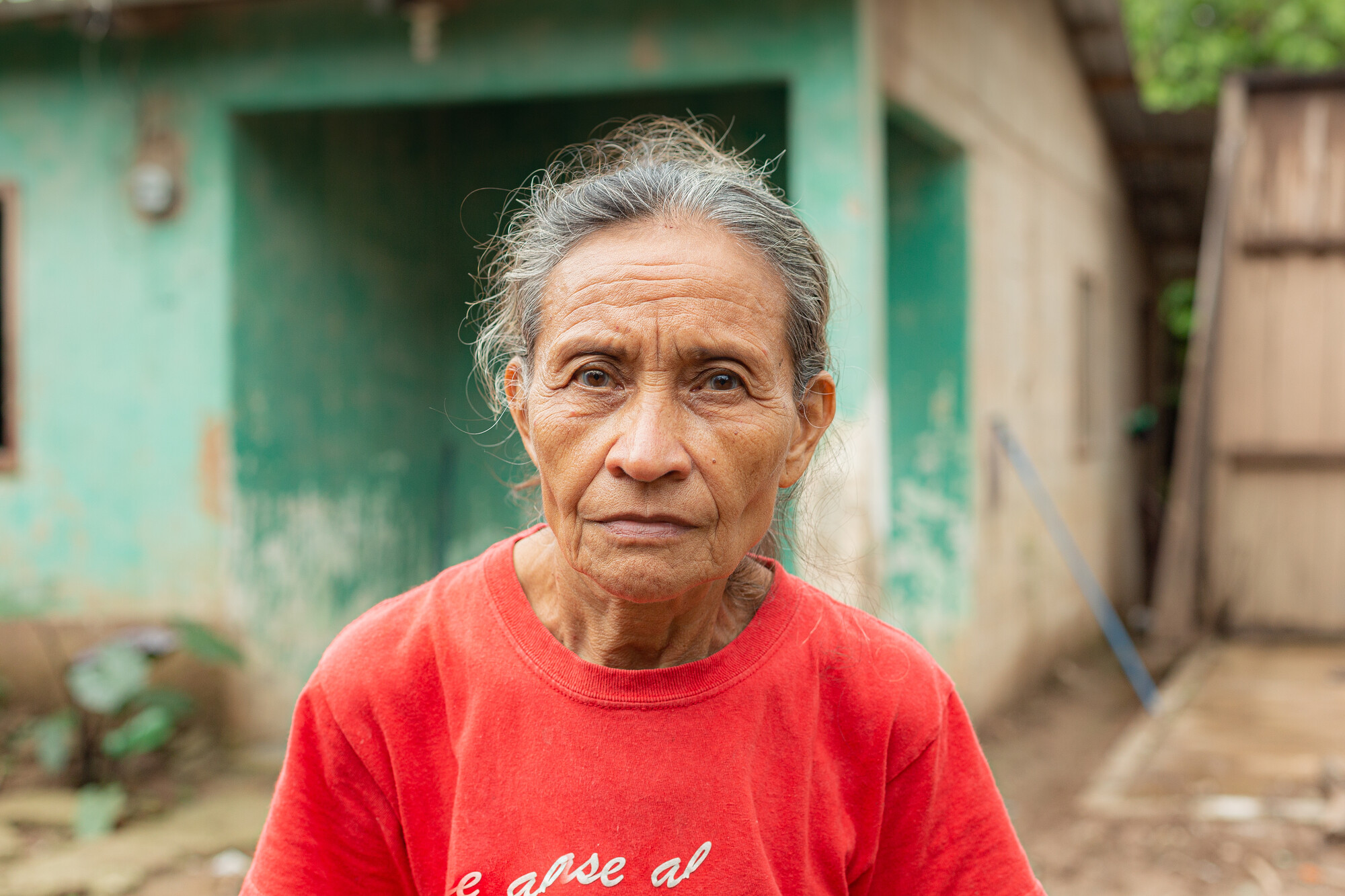 A older Honduran woman in a red shirt stands in front of a house