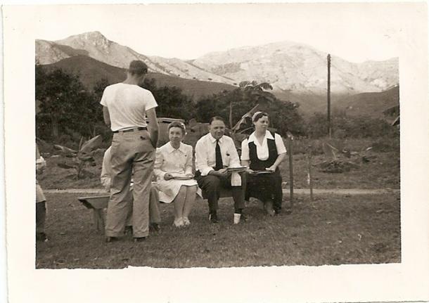 A group of three people sitting on a bench next to a man who is standing next to them with his back to the camera