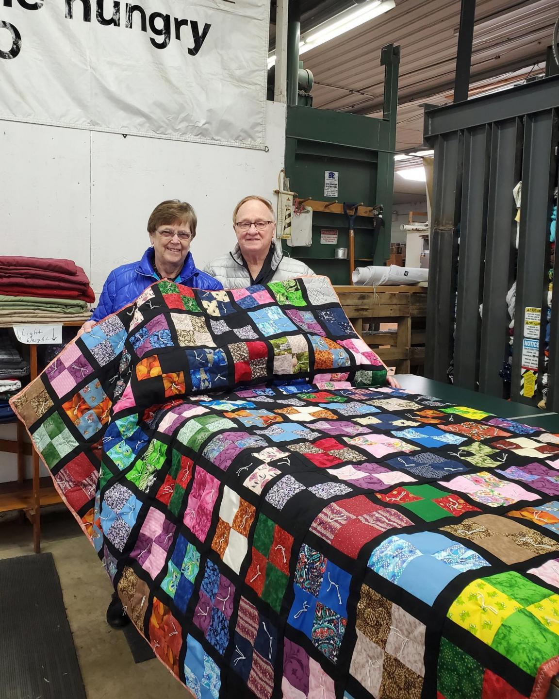 Two people display a colorful comforter on a table in front of them in a warehouse.