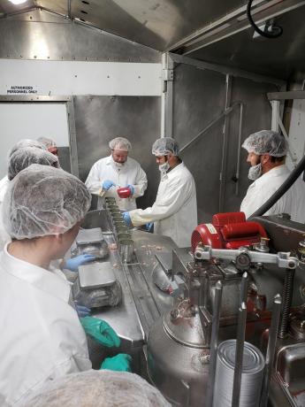 A group of people in white coats and hair nets work on a canning conveyer line