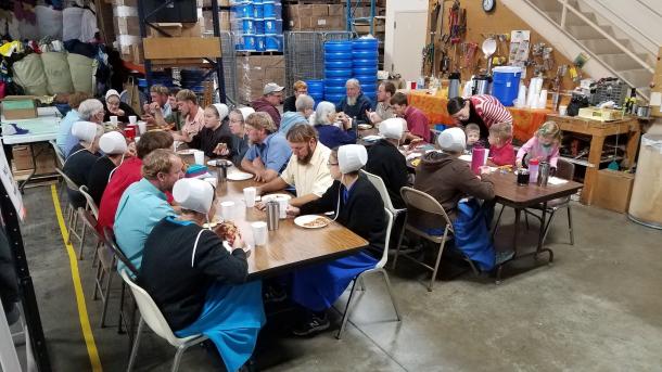 A large group of people sit at tables and eat. They are in a warehouse