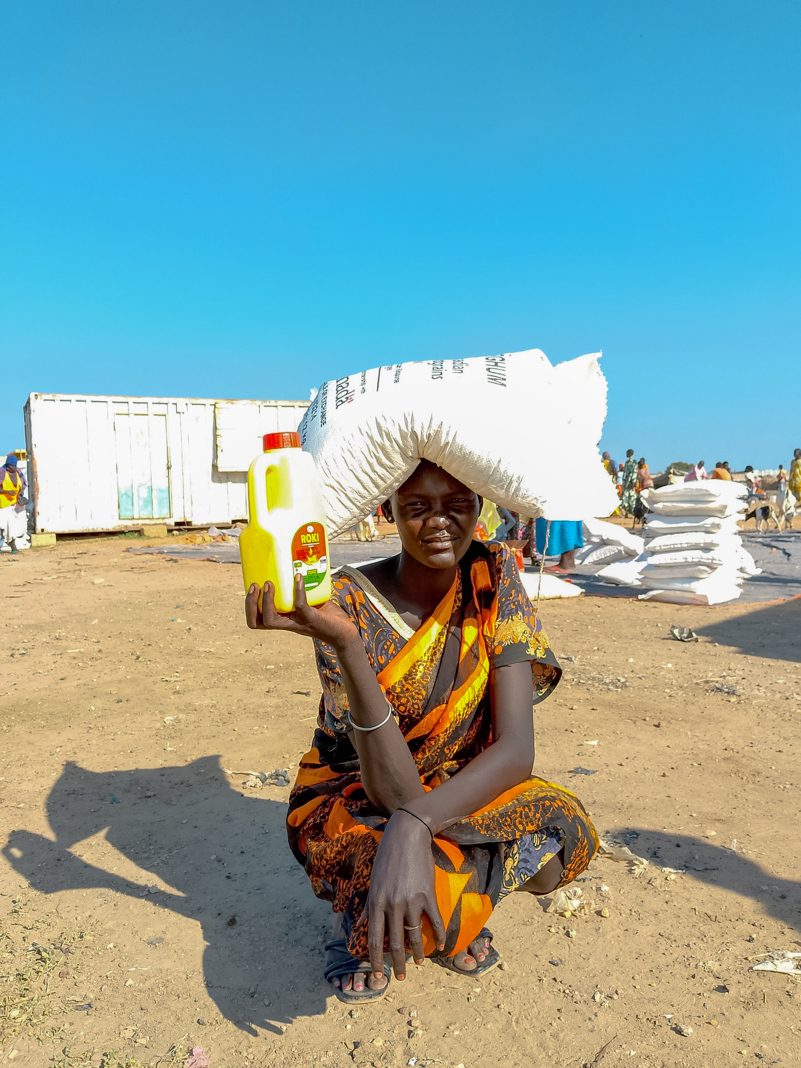 A woman from South Sudan named Martha squats in an open area next to a food distribution. She's holding food she recently received.