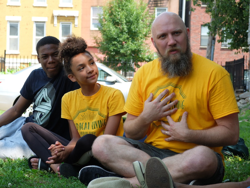 Chloe Storbakken and Jason Storbakken sit as part of a circle on the grass in Brooklyn, New York's Bedford-Stuyvesant neighborhood in July 2019 with Radical Living youth summer camp participants. They