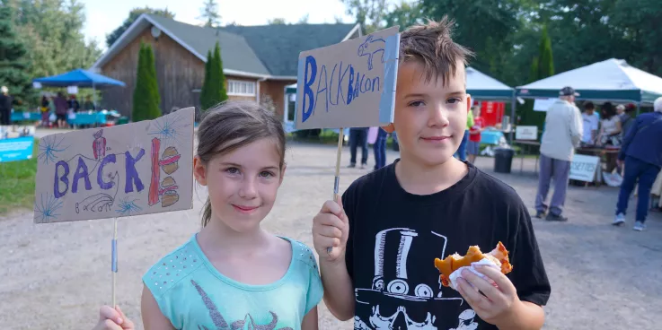 A boy and a girl hold up hand made cardboard signs that read Back Bacon, while the boy eats bacon on a bun.