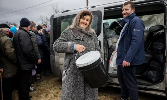 A woman in a winter coat holds up a five gallon bucket and smiles at the camera.