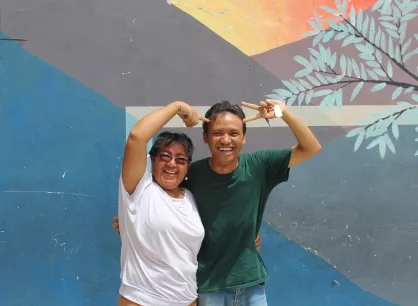 Two people stand with their arms around each other. One is pointing at the other and the other is doing a peace sign.