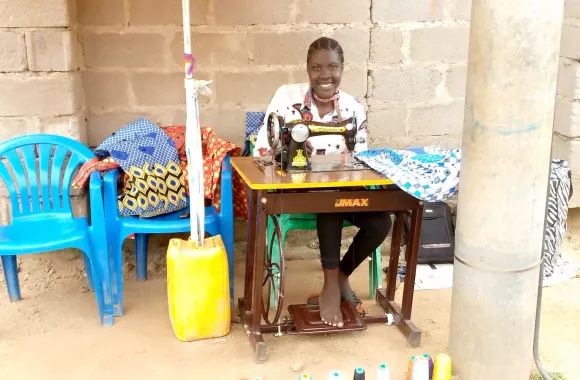 A person sits at a sewing machine and smiles at the camera