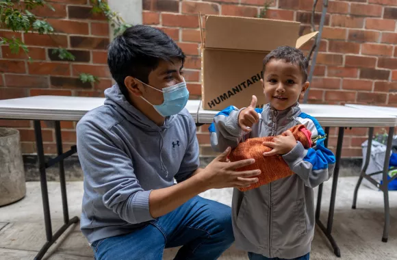 A man crouches down near a boy holding an orange bag and giving a thumbs up.