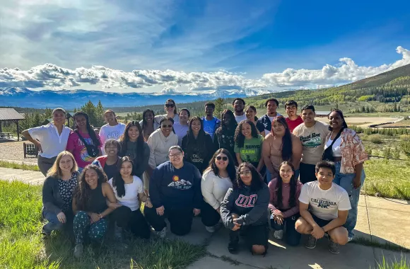 A large group of young adults pose for a photo. In the background is a mountain range.