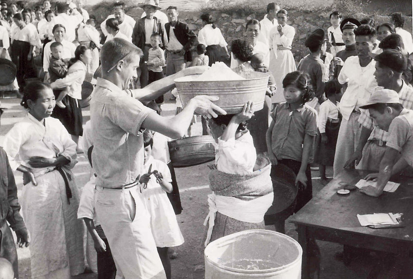 A black and white photo from the 1950s of a food distribution in Korea. A man is placing a large bucket of grain on a person's head.