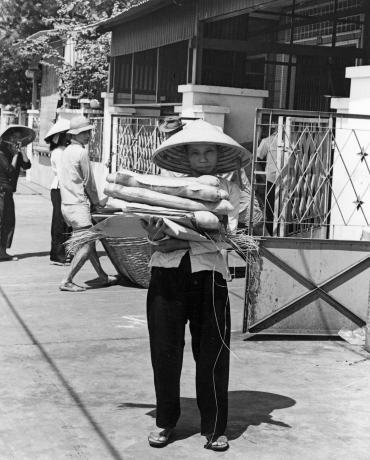 A black and white photo from the 1960s of a person in Vietnam carrying a pile of bread loaves