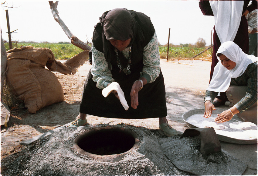 A Jordanian woman leans over an in-ground oven and shapes flat bread dough