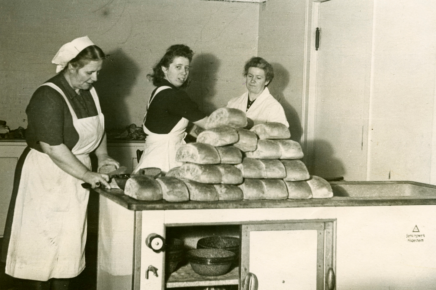A black and white photo from teh 1940s of three women working with a large pile of bread loaves