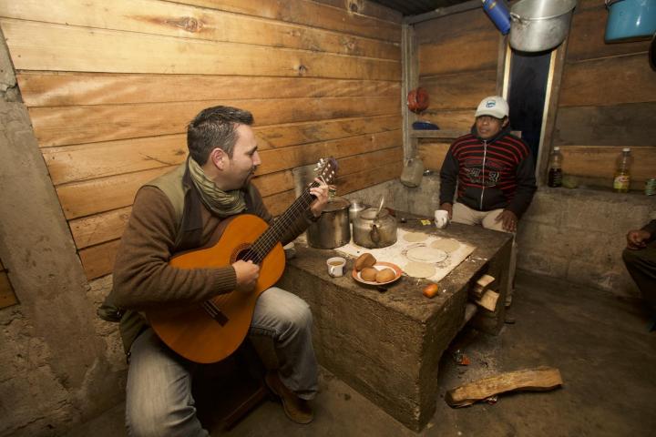 Two men sit at a small table. One man is playing the guitar