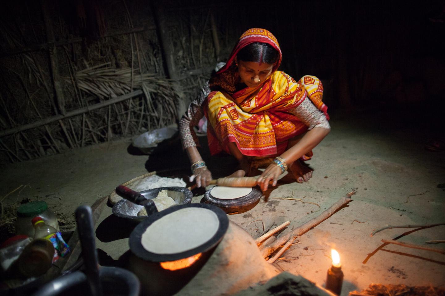 A Nepali woman squats on the ground and rolls out flat bread by her outdoor tandoori oven.She is working by candlelight