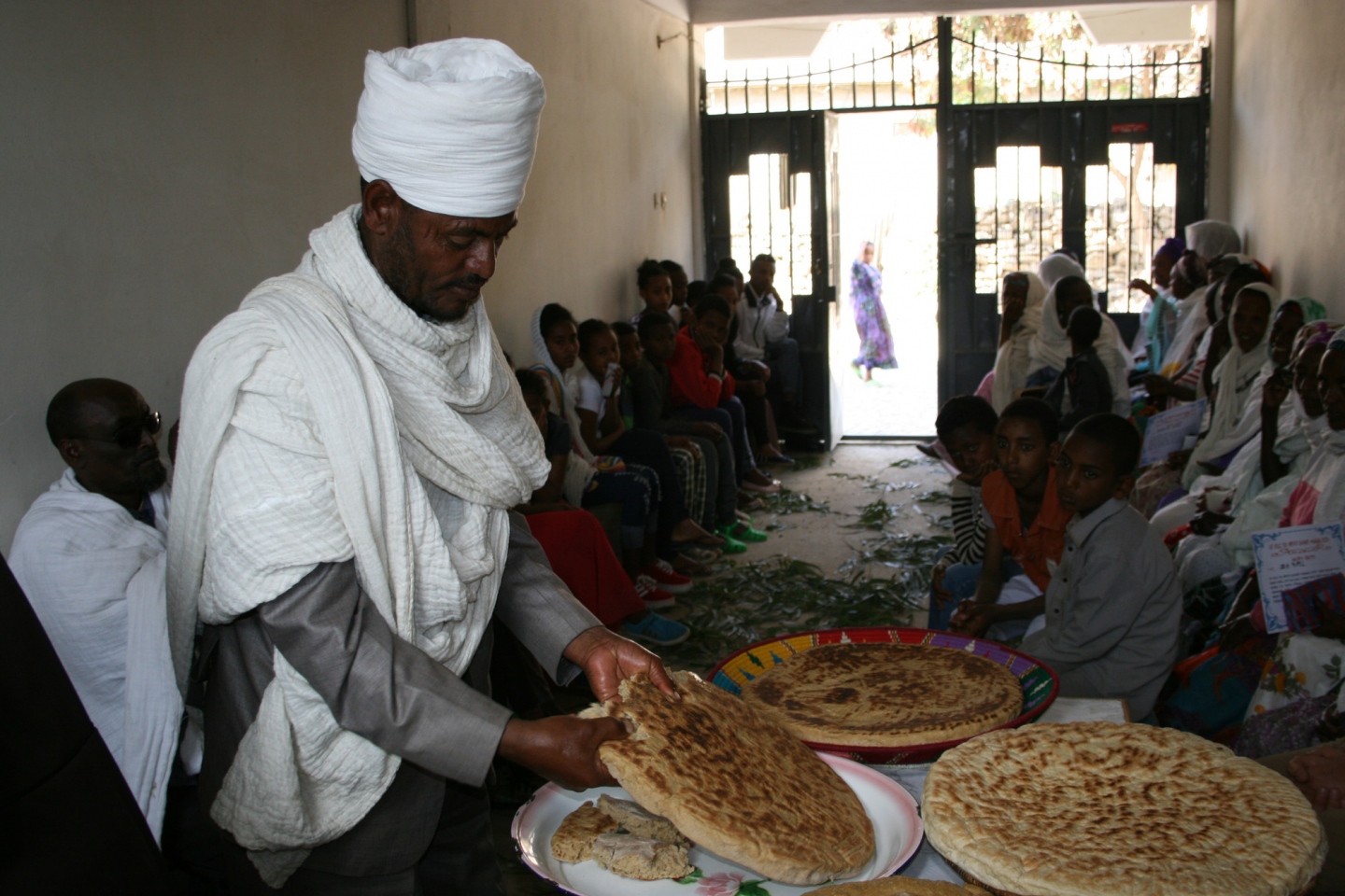 An Ethiopian man in a white turban and white scarf tears off a pieace of bread. He is in a room with a large group of people.