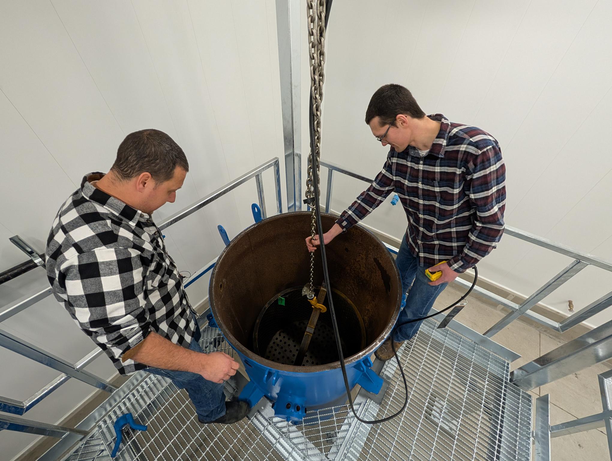Two men look into a large round metal container.